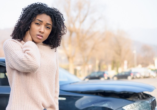 A young Ghanaian woman holds her neck in the aftermath of a car accident. A woman holds her neck and stands next to her car after being involved in an accident.