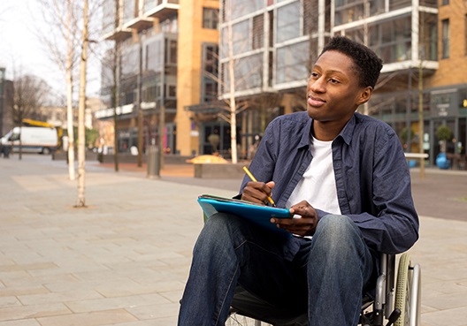 A young man sits in a wheelchair, having become disabled after an accident. A young man seated in a wheelchair writes down notes outdoors.