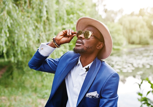 A man wearing a blue blazer looks up at the sky through sunglasses. A Ghanaian man wearing sunglasses looks up towards the sun.