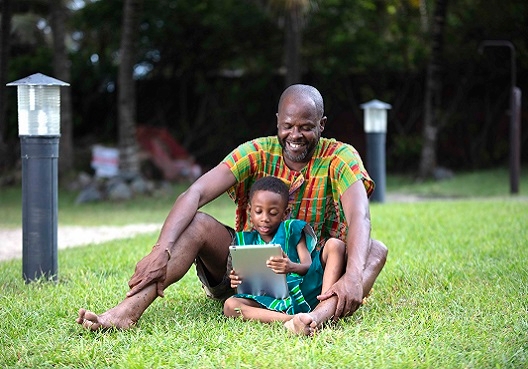 A man enjoys sitting and reading with his son, resting easy knowing he is investing in his education. A Ghanaian father and son sit outdoors and read together.
