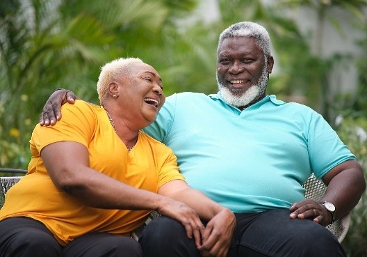 An elderly couple smile and laugh as they spend time together in their retirement. An older couple sit on a bench and enjoy the outdoors together.
